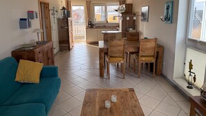 Amplia sala de estar con sofá, mesa de centro y mesa de comedor de madera con sillas. Se ve una cocina al fondo, con una ventana y vistas al exterior.
