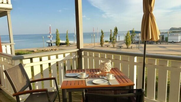 A wooden deck with a table set for two, overlooking a body of water and a marina.