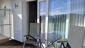 Un balcón con dos sillas, una mesa, un ventanal corredero y una pared blanca con una ventana.