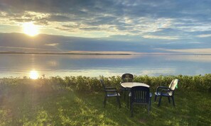 Un espace herbeux avec une table et des chaises, donnant sur une étendue d’eau calme sous un ciel nuageux.