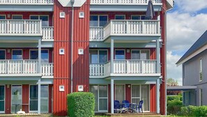 A red and white multi-story building with balconies, a small garden, and outdoor seating. - Mitteldeck 464 - Gemütliches Ferienapartment mit Blick auf die Müritz (Rechlin-Nord)