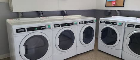 Une rangée de machines à laver Maytag à chargement frontal, toutes blanches, dans une laverie, avec une horloge et des panneaux indiquant les services de blanchisserie au-dessus.
