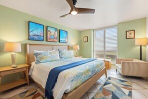 A bedroom with a bed, two bedside tables, a ceiling fan, a window with blinds, and a chair. - D27 North Coast Village - Oceanfront, ground level cottage, Amazing Direct Ocean Views (Oceanside)
