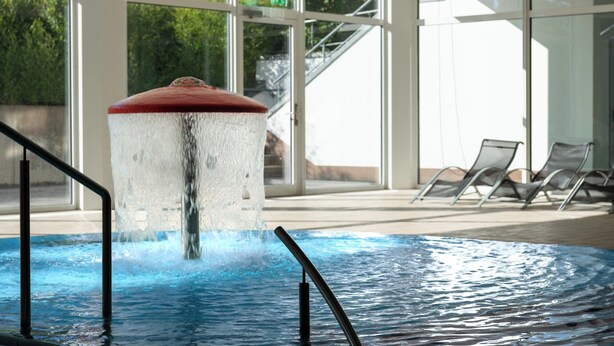 A swimming pool with a water feature, indoor seating, and large windows.