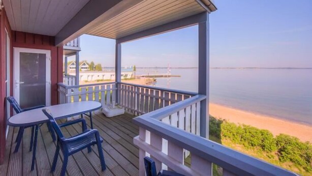 A wooden deck with a table and chairs, overlooking a body of water and a distant shoreline.