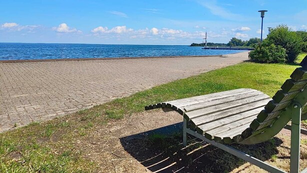 A beach with a paved walkway, a wooden bench, and a lamp post. - Mitteldeck 504 - Cozy vacation apartment with a view of the Müritz (Rechlin-Nord)