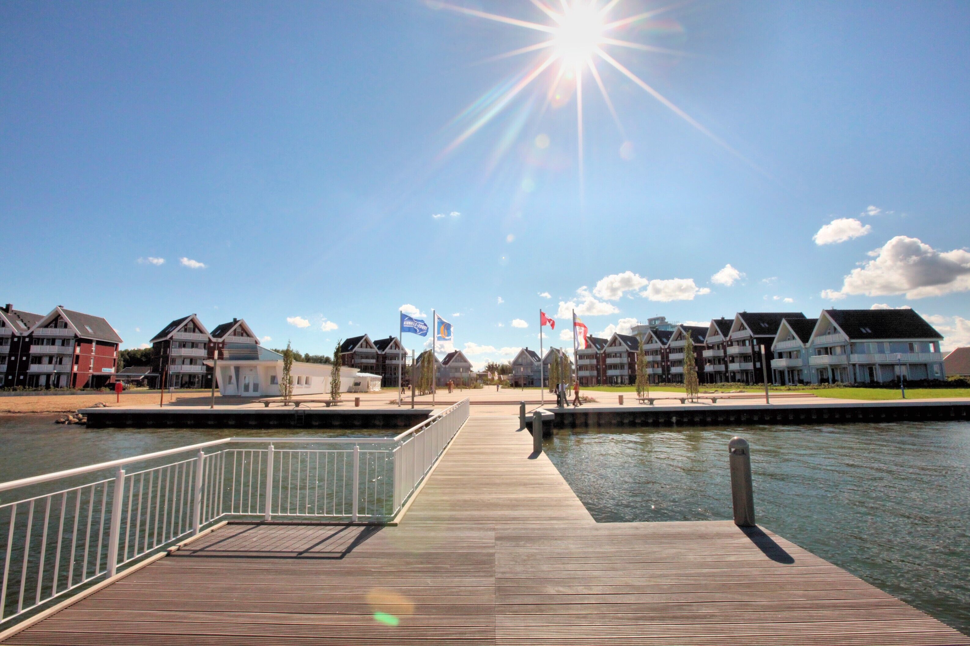 A wooden pier extending over calm water, leading to a row of modern houses with flags.