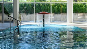 Ein Schwimmbecken mit zentralem Wasserspiel, umgeben von großen Fenstern und einer Betonmauer.