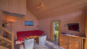 Un acogedor interior de cabaña con un sofá rojo, dos sillones blancos, una mesa de comedor de madera, un televisor montado en la pared y un aparador de madera.