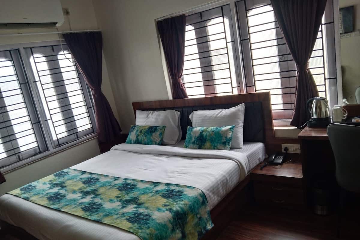 A bedroom with a bed, two pillows, a bedside table with a kettle and a phone, and windows with curtains.