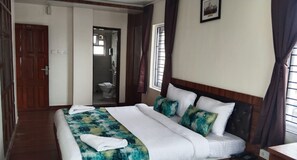 Une chambre d’hôtel avec un grand lit, une porte en bois, une salle de bain visible à travers une porte ouverte et un tableau encadré au mur.