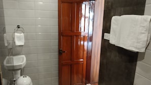 A bathroom with a wooden door, white tiles, a sink, and a towel hanging on the wall. - Hotel Kolkata (Barasat)