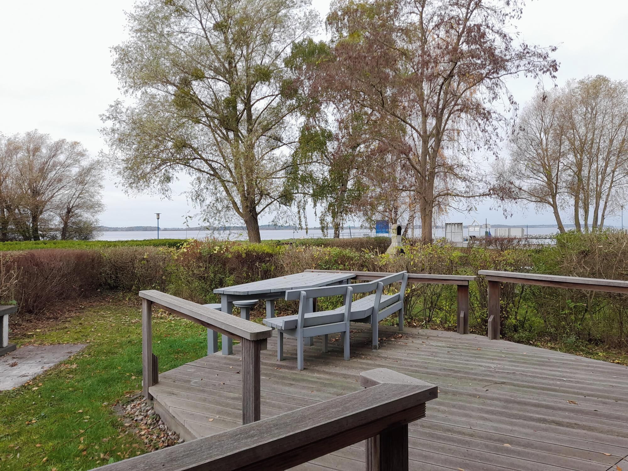 A wooden deck with a table and chairs, overlooking a body of water with trees in the background.