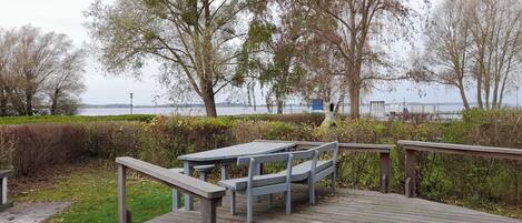A wooden deck with a table and chairs, overlooking a body of water with trees in the background.