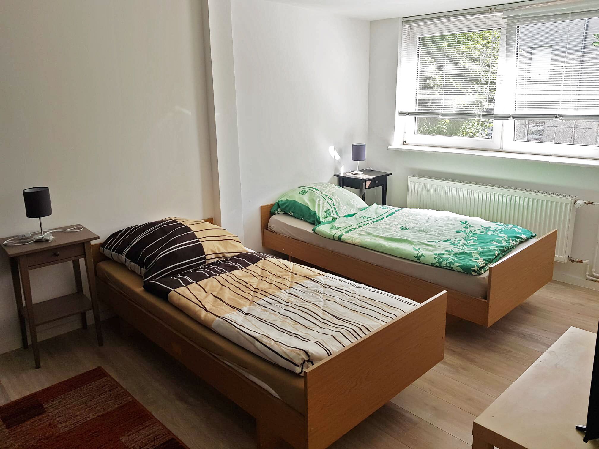 A bedroom with two wooden beds, a nightstand with a lamp, a radiator, and a window with blinds.