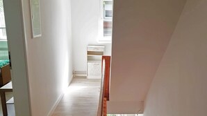 Um corredor amplo com piso de madeira, parede branca e janela com persianas.