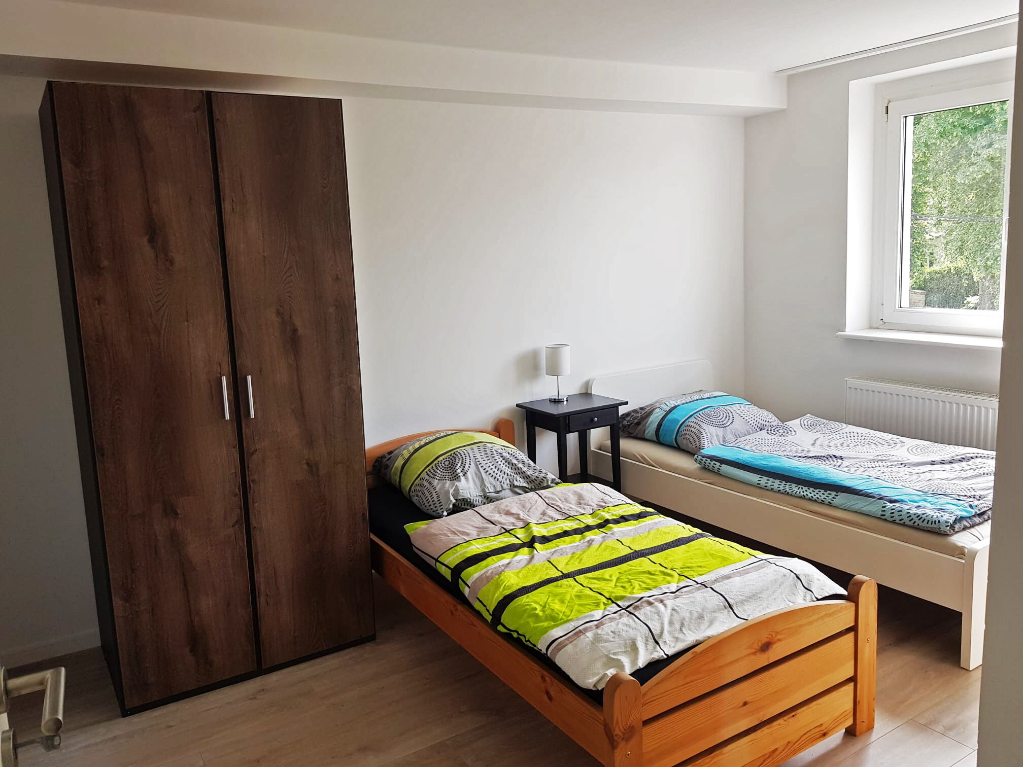 A bedroom with a wooden wardrobe, a single bed with a bright green and white duvet, a bedside table with a lamp, and a window with a view of greenery.