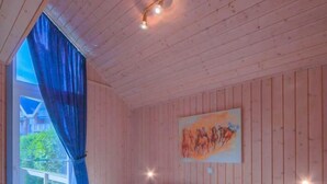 A wooden cabin bedroom with a bed, a nightstand, a window with curtains, and a painting on the wall.