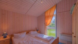 A wooden cabin bedroom with two beds, a nightstand with a lamp, and a window with curtains.
