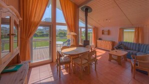 A cozy room with a wooden ceiling, a round dining table with chairs, a sofa, and a view of a green landscape through large windows.