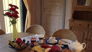 Una mesa de comedor de madera dispuesta con platos, tazones, tazas y una tetera, rodeada de sillas en una habitación con paredes de madera y una ventana con cortinas.