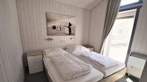 Une chambre à coucher avec un lit en bois, une table de chevet, un petit tiroir, un tableau accroché au mur et une fenêtre avec des rideaux.