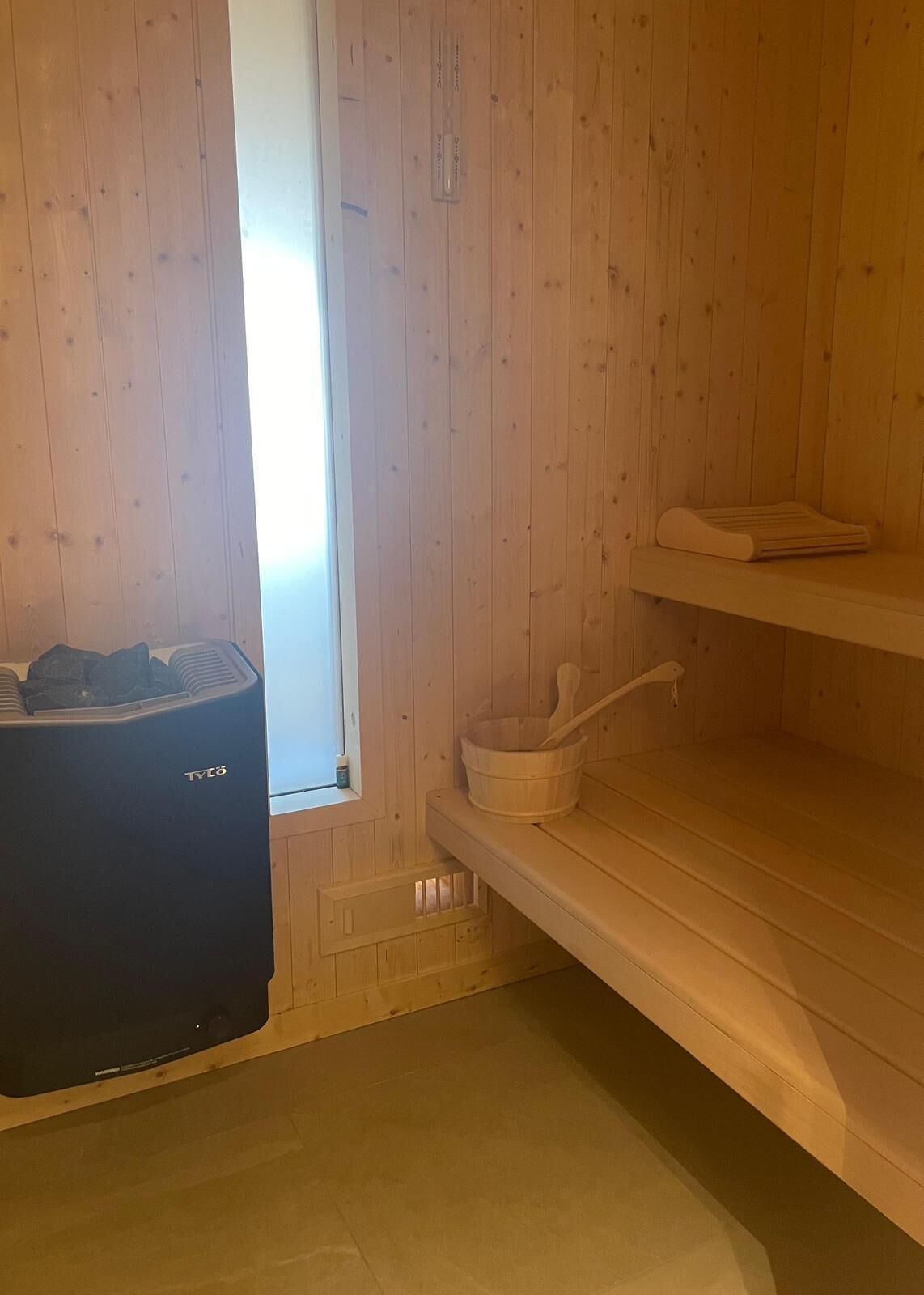 A sauna room with wooden walls, a black sauna heater, a wooden bucket, and a towel on a shelf.