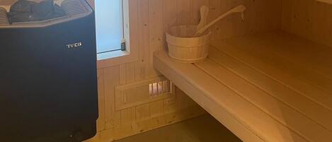 A sauna room with wooden walls, a black sauna heater, a wooden bucket, and a towel on a shelf.