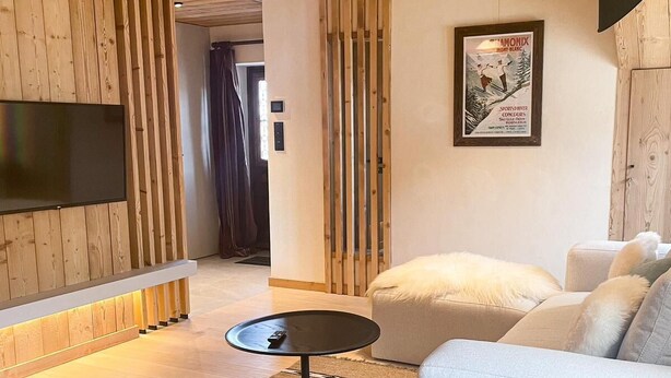 Ein modernes Wohnzimmer mit Holzboden, einem großen Flachbildfernseher an der Wand, einer Couch, einem Couchtisch und einem Holzstuhl.