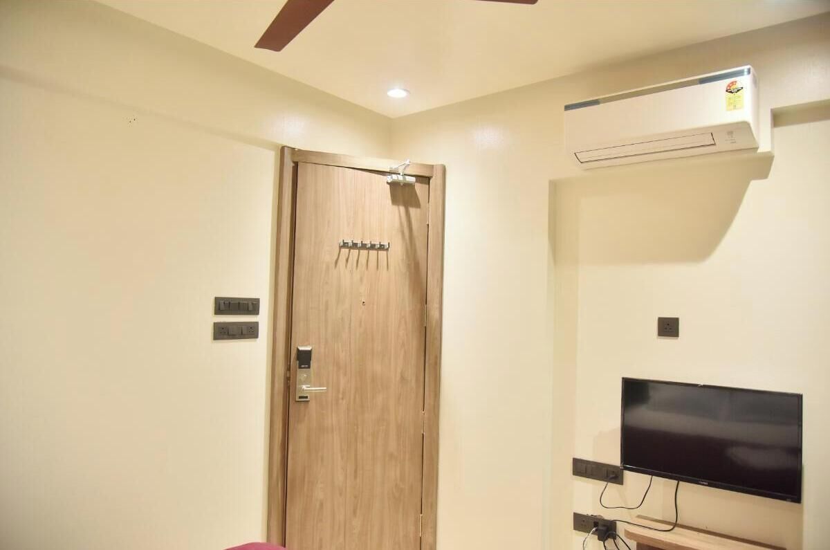 A room with a wooden door, an air conditioning unit, a flat-screen TV mounted on the wall, and a ceiling fan.
