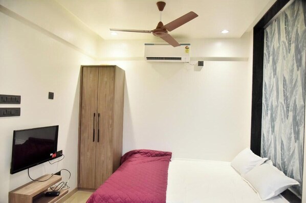 A bedroom with a bed, a wardrobe, a ceiling fan, an air conditioner, a TV, and a window with curtains.