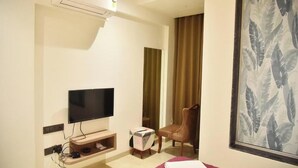 A hotel room with a flat-screen TV mounted on the wall, a wooden chair, a small table with a remote, and a large window with a tropical leaf pattern. - Hotel Manoranjan Enclave (Dindori)