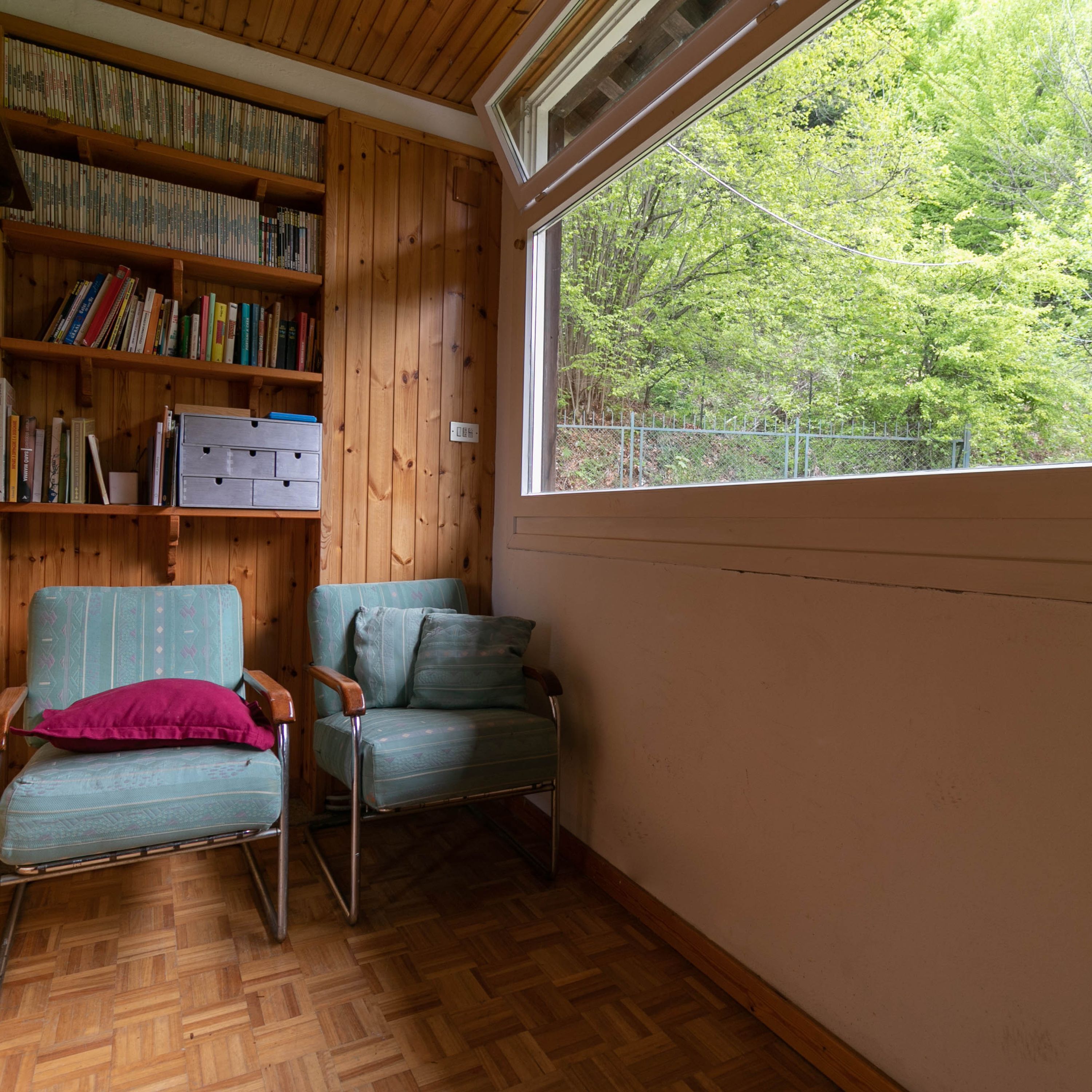 A room with a wooden bookshelf, two chairs, a window with a view of trees, and a herringbone-patterned floor.