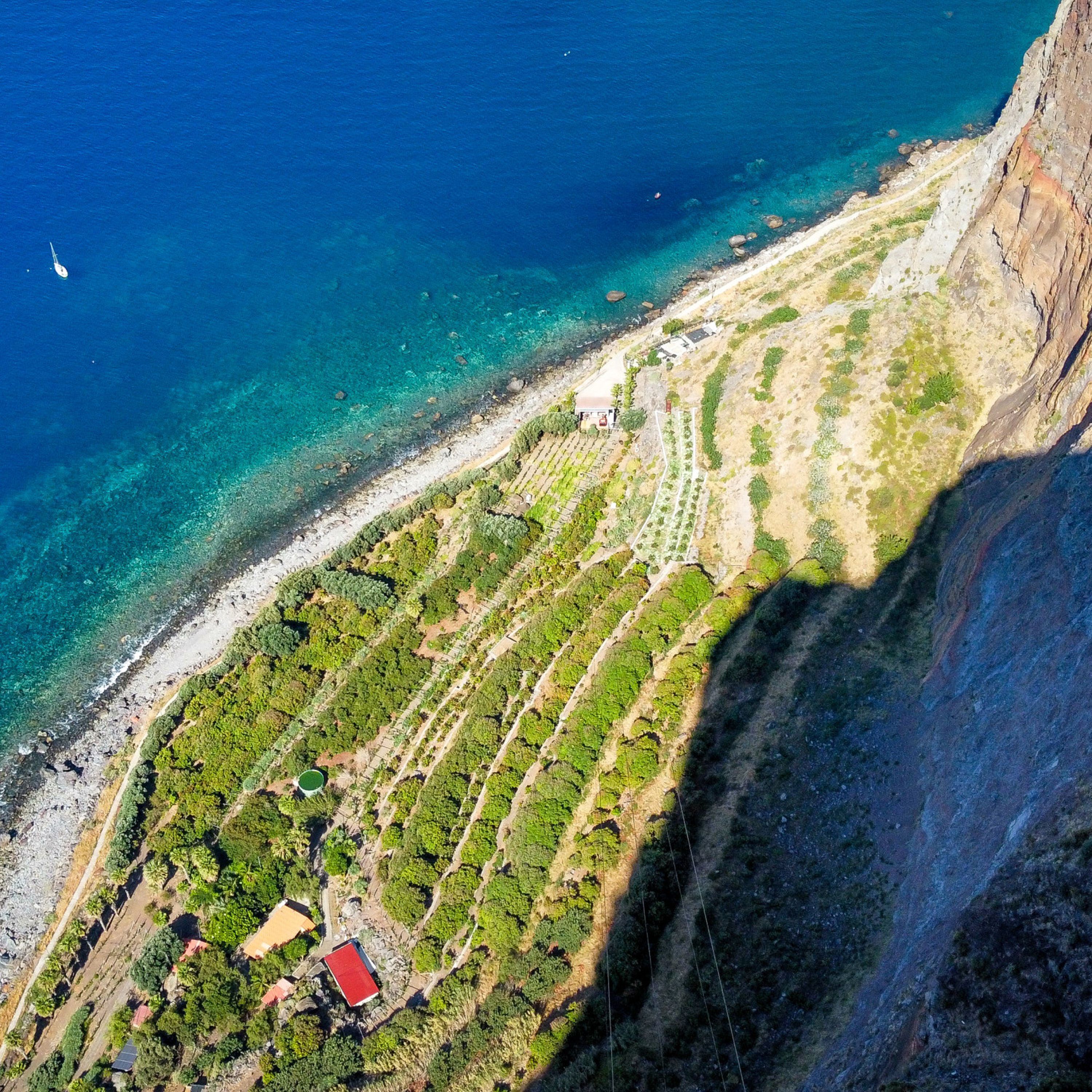 An aerial view of a coastal area with terraced fields, a small building with a red roof, and a cliffside.