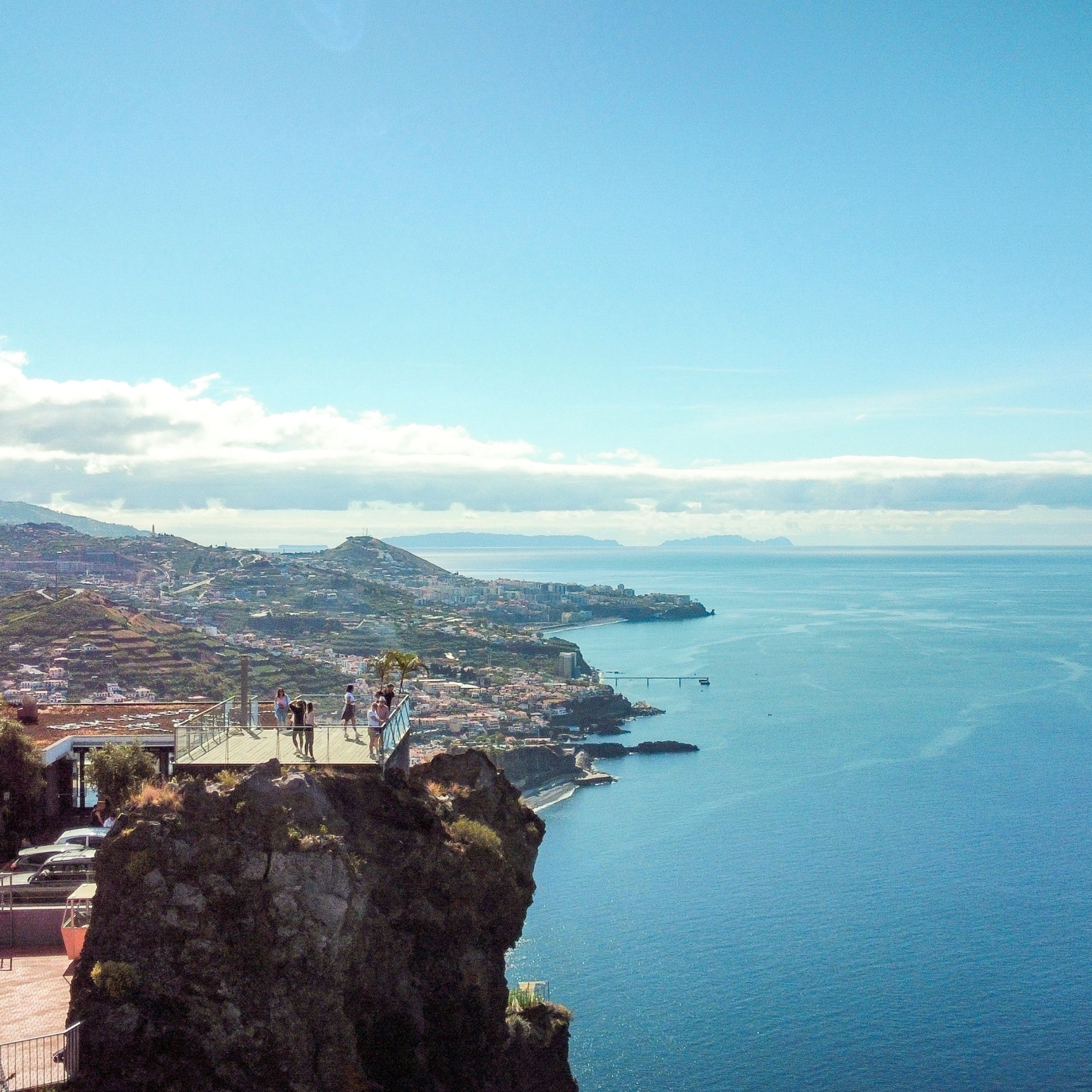 A coastal view with a rocky outcrop and a viewing platform overlooking the sea and a distant cityscape.