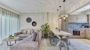 A modern living room with a sofa, a dining table, and bar stools. There is a kitchen area with a refrigerator, microwave, and oven.