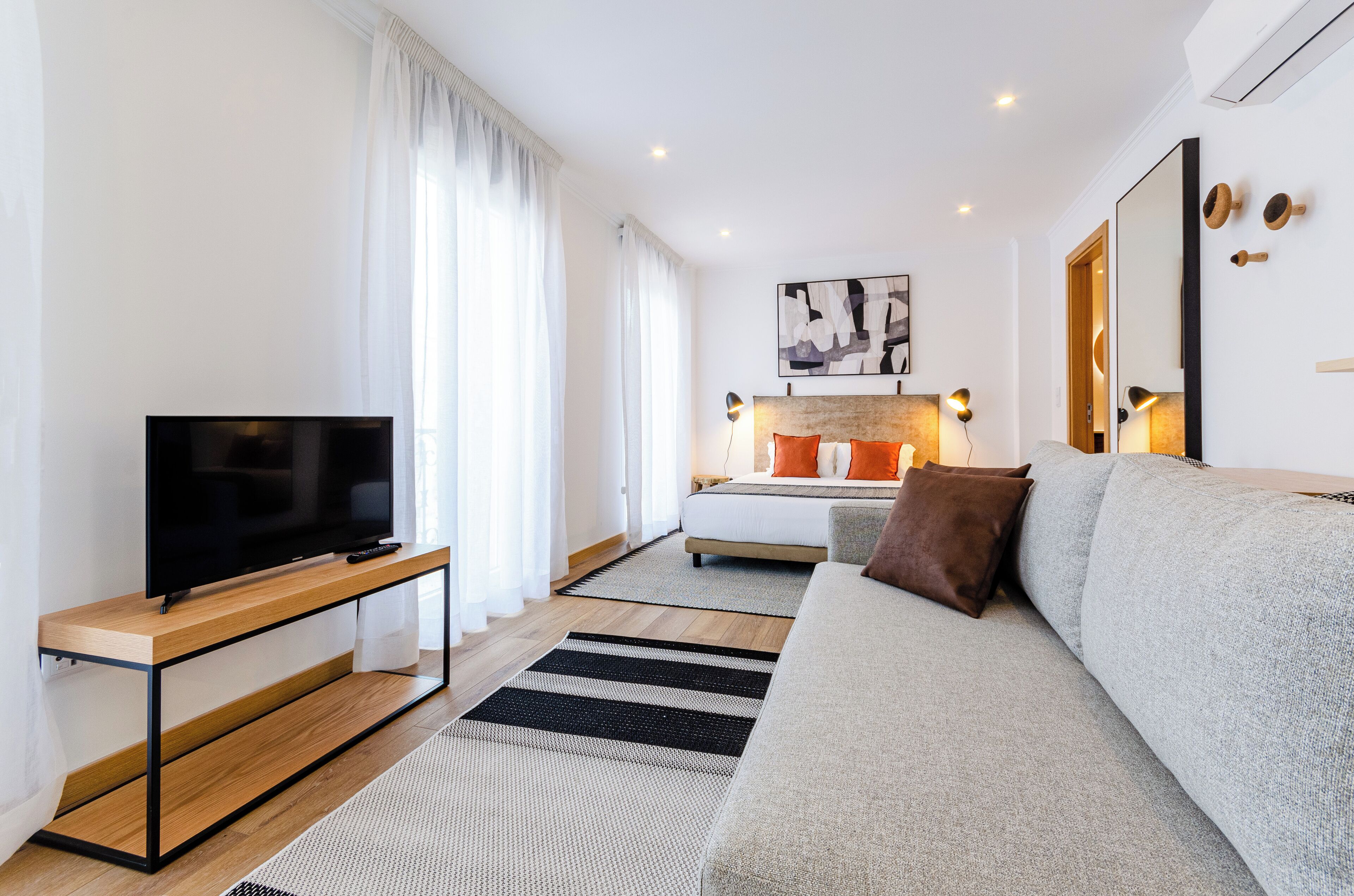 Una sala de estar moderna con una televisión de pantalla plana sobre una consola de madera, un sofá gris y una cama con almohadones naranjas.
