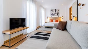 Una sala de estar moderna con una televisión de pantalla plana sobre una consola de madera, un sofá gris y una cama con almohadones naranjas.