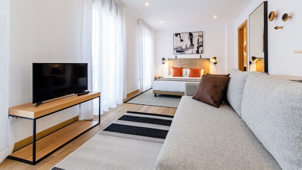A modern living room with a flat-screen TV on a wooden console, a grey sofa, and a bed with orange pillows. - Morais Soares 131 (Lisbon)