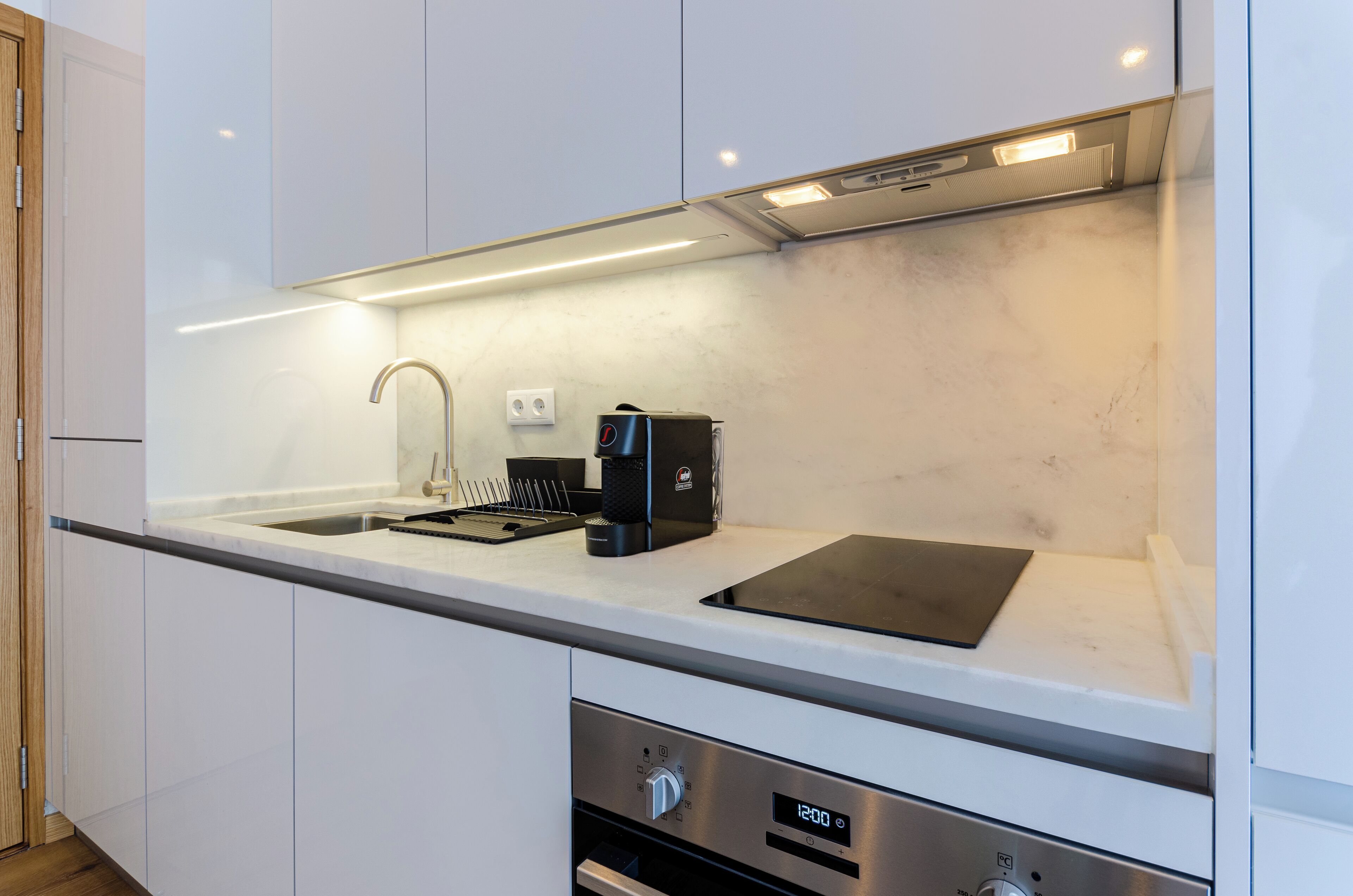 Una cocina moderna con un lavamanos de acero inoxidable, una cafetera negra y un reloj digital que muestra las 12:00.