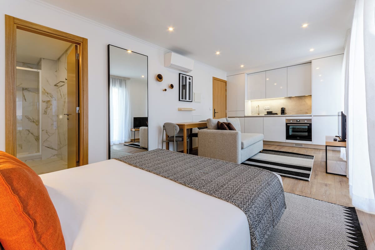 A modern hotel room with a large bed, a sitting area with a table and chairs, a kitchenette, and a bathroom with a glass shower.