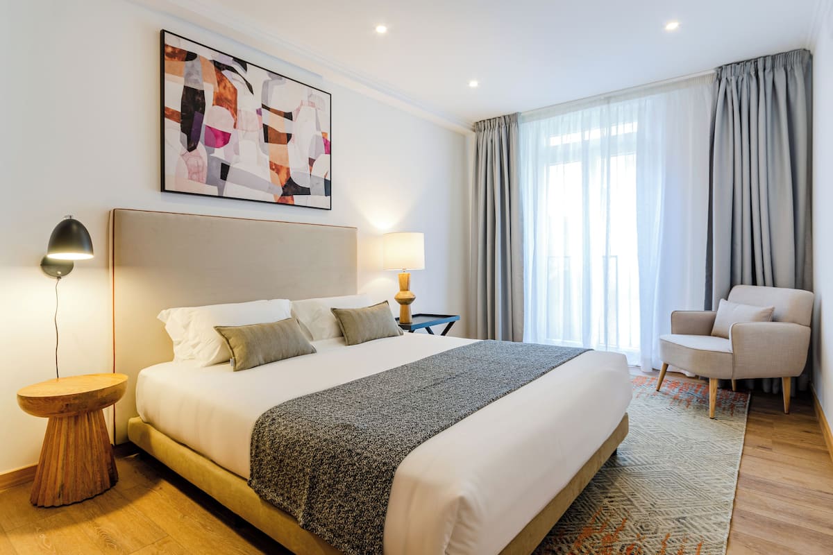 A hotel room with a large bed, a side table, a lamp, a chair, and a window with curtains.