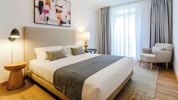 Habitación de hotel con una cama grande, una mesita de noche, una lámpara, una silla y un ventanal con cortinas.