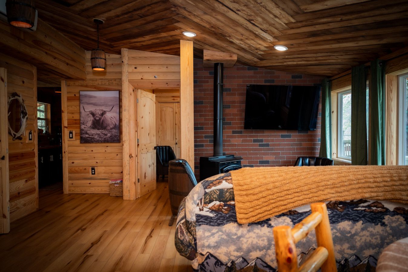 A log cabin interior with a wooden floor, a bed with a patterned blanket, a mounted television, and a fireplace.