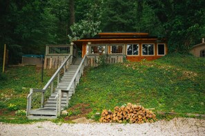 A cabin with a wooden exterior, a staircase leading to a deck, and a pile of firewood in front.