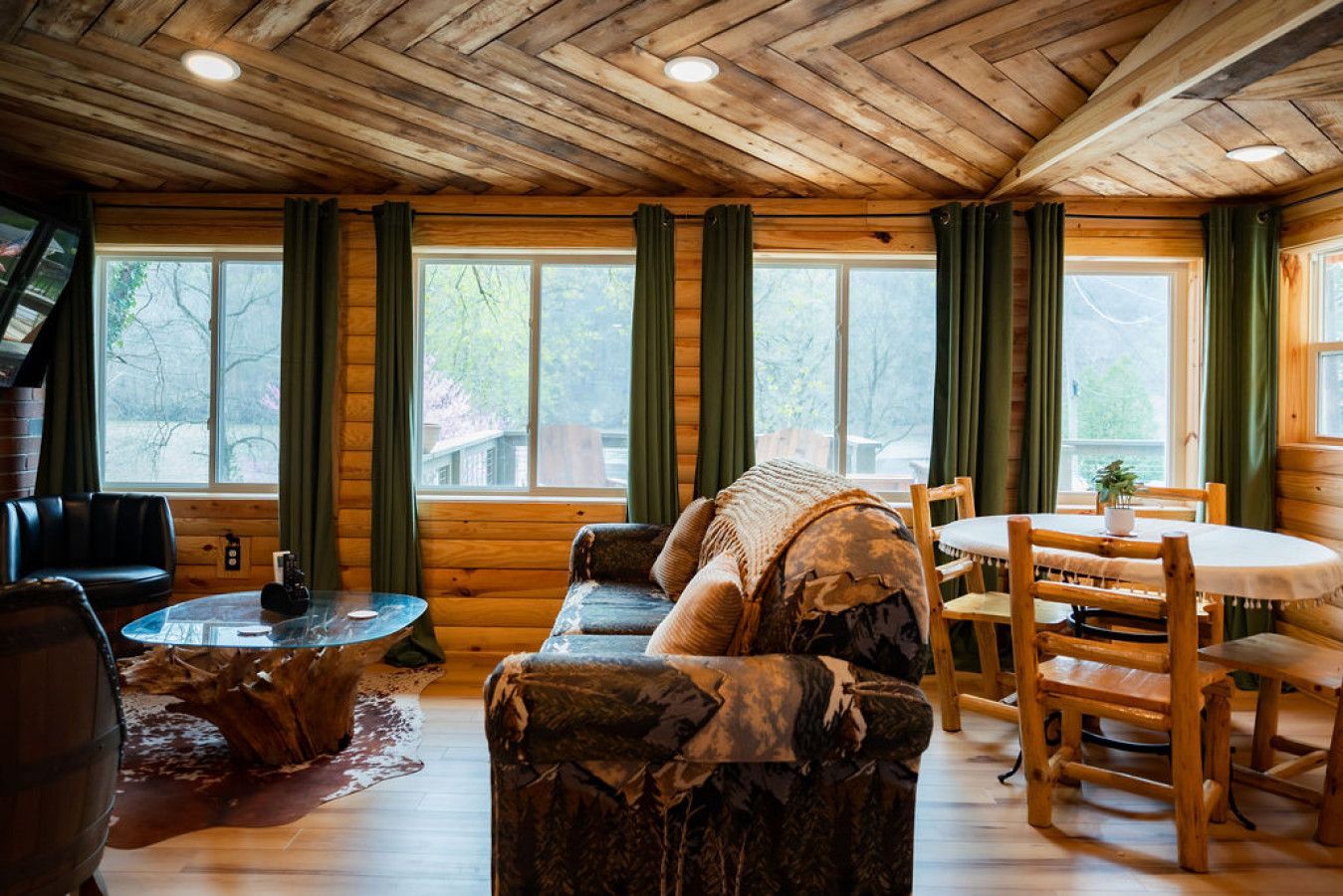 L’intérieur d’un chalet accueillant, avec un plafond en bois, de grandes fenêtres dotées de rideaux, un canapé, une table à manger avec des chaises et une petite table ronde sur laquelle se trouve une bouteille.