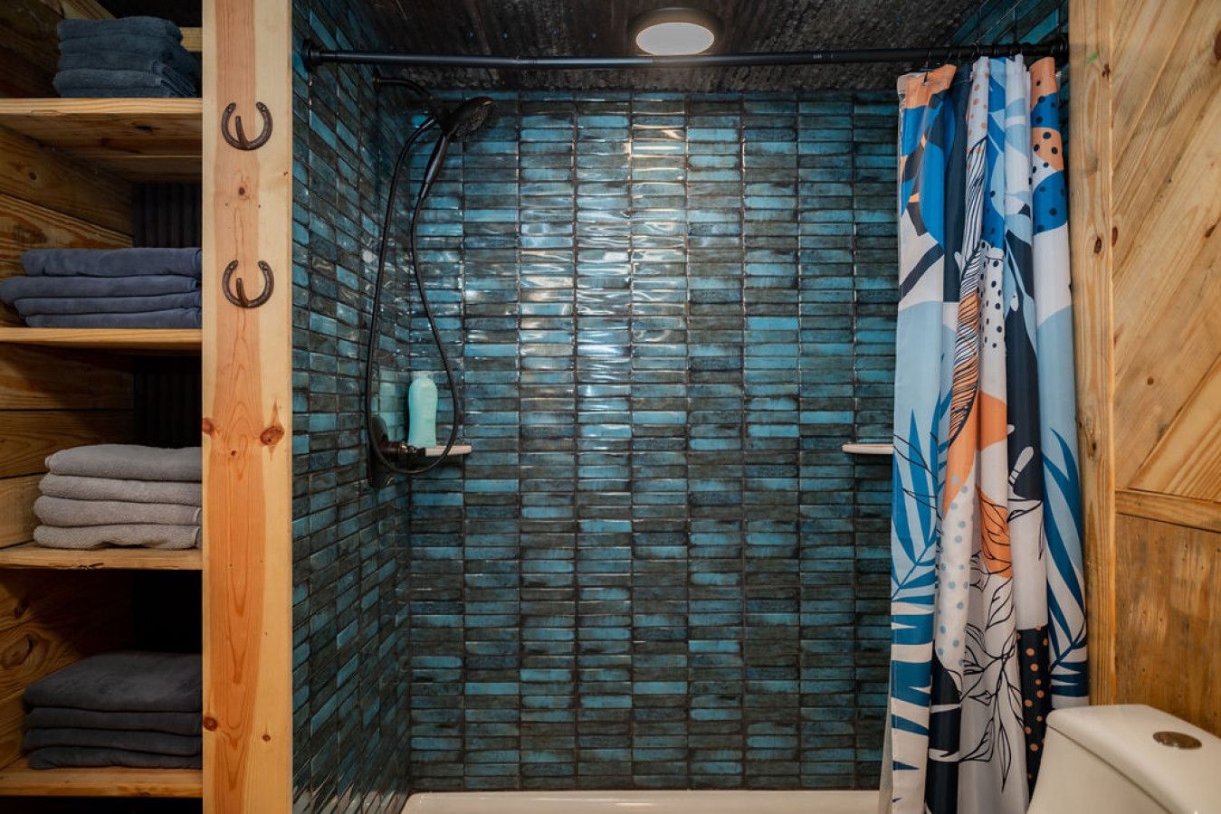 Une douche avec un mur recouvert de carreaux bleus, des étagères en bois où sont pliées des serviettes, et un rideau de douche à motifs.