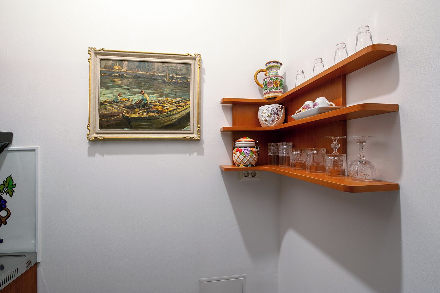 A room with a framed painting of a boat on water, wooden shelves with glassware and decorative items, and a white wall.