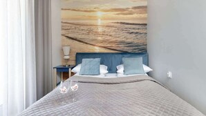 Una habitación de hotel con cama, mesita de noche, lámpara y un mural en la pared que muestra un atardecer sobre el océano.