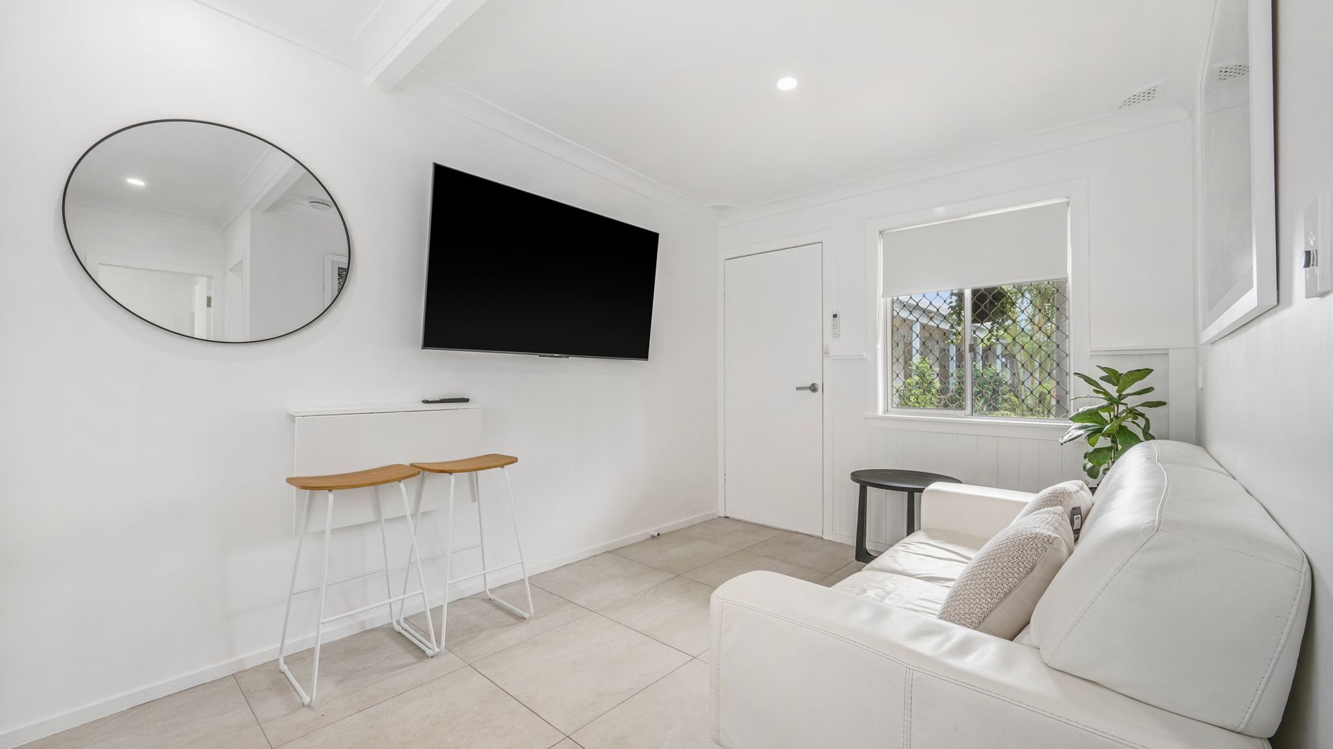 Un salón moderno con un sofá blanco, un televisor montado en la pared, un espejo redondo y una pequeña mesa con taburetes.